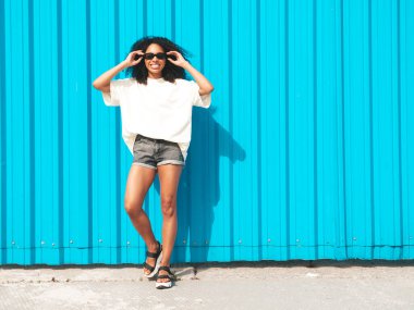 Saçları kıvırcık, güzel siyah bir kadın. Beyaz tişörtlü, hippi bir model. Sokakta mavi duvarın yanında güneş gözlükleriyle poz veren kaygısız seksi kadın. Neşeli ve mutlu