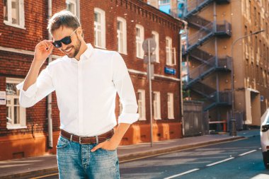 Yakışıklı, gülümseyen stil sahibi Lambersex modelinin portresi. Beyaz gömlek giymiş modern bir adam. Sokakta güneş gözlükleriyle poz veren modacı erkek. Gün batımında dışarıda. 