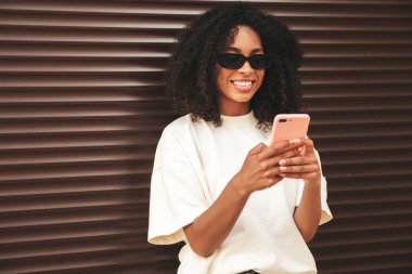 Saçları kıvırcık, güzel siyah bir kadın. Beyaz tişörtlü, hippi bir model. Sokakta güneş gözlüklü, kahverengi duvarın yanında poz veren seksi kadın. Akıllı telefon ekranına bakıyor, uygulamalar kullanıyor.