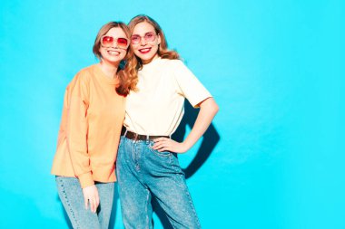 İki genç, güzel, gülümseyen yeniyetme kadın, modaya uygun yaz hippi kıyafetleri içinde. Stüdyoda mavi duvarın yanında poz veren kaygısız seksi kadınlar. Moda ve pozitif modeller eğleniyor. Neşeli ve mutlu