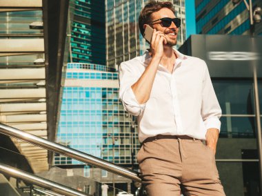 Yakışıklı, güler yüzlü, stil sahibi, lamberseksüel model. Beyaz gömlekli, modern bir adam. Sokakta güneş gözlükleriyle poz veren modacı erkek. Gün batımında dışarıda. Akıllı telefon kullanarak. Konuşarak.