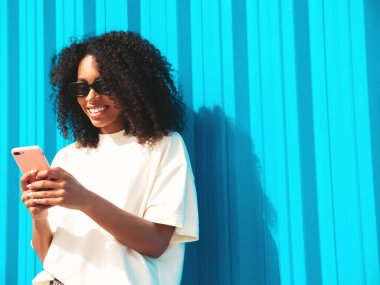 Saçları kıvırcık, güzel siyah bir kadın. Beyaz tişörtlü, hippi bir model. Sokakta güneş gözlüklü, mavi duvarın yanında poz veren seksi kadın. Akıllı telefon ekranına bakıyor, uygulamalar kullanıyor.