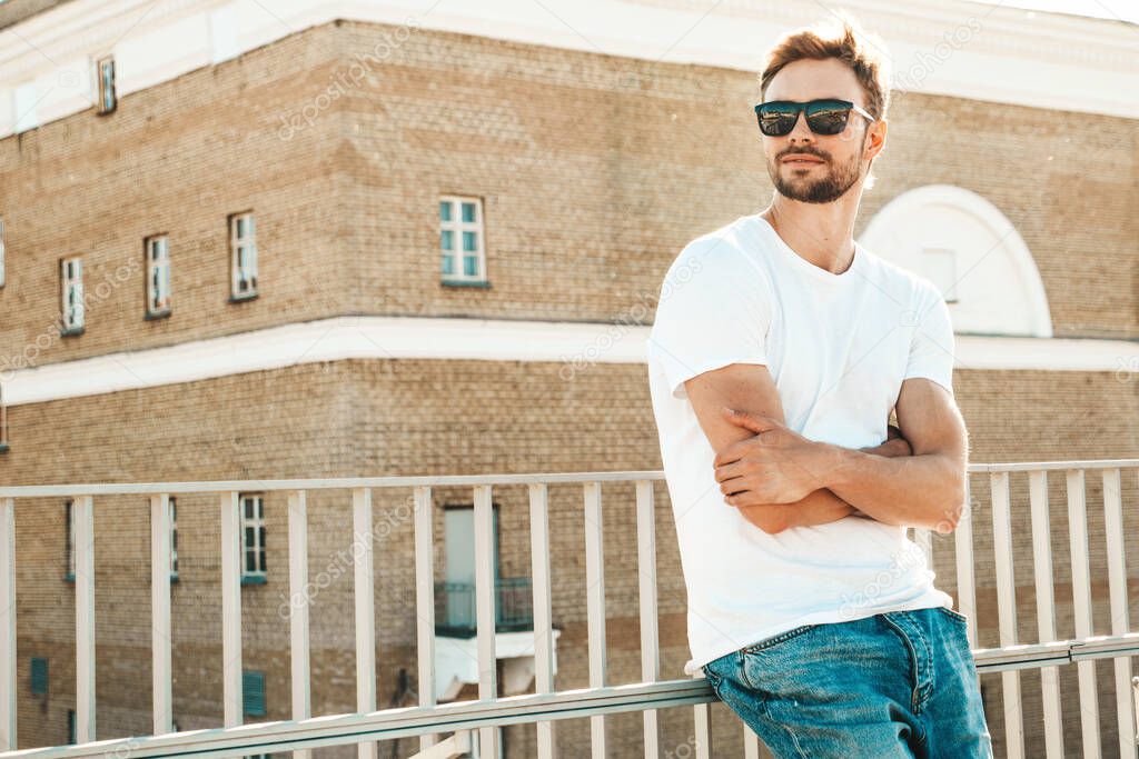 Portrait of handsome smiling  hipster lambersexual model.Stylish man dressed in white T-shirt and jeans. Fashion male posing on the street background near fence at the beach 