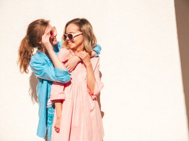 Son moda yaz kıyafetleri ve elbiseleri içinde iki güzel, gülümseyen hippi kadın. Sokakta beyaz duvarın yanında poz veren kaygısız kadınlar. Pozitif saf modeller eğleniyor, sarılıyor ve deliriyor.