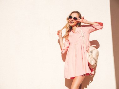 Son moda pembe elbiseli güzel, gülümseyen hippi kadın. Günbatımında sokakta beyaz duvarın yanında poz veren kaygısız seksi kadın. Olumlu model açık havada. Mutlu ve neşeli. Barış işareti nasıl?