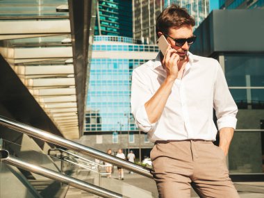 Yakışıklı, güler yüzlü, stil sahibi, lamberseksüel model. Beyaz gömlekli, modern bir adam. Sokakta güneş gözlükleriyle poz veren modacı erkek. Gün batımında dışarıda. Akıllı telefon kullanarak. Konuşarak.