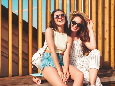 Son moda yaz kıyafetleri içinde iki güzel gülümseyen yeniyetme kadın. Sokakta şapkalı kayla poz veren kaygısız kadınlar. Pozitif saf modeller gün batımında eğleniyorlar, sarılıyorlar ve deliriyorlar.