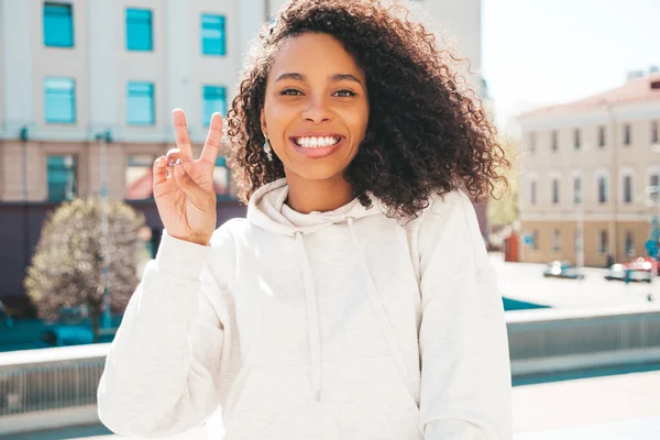 Beautiful black woman with afro curls hairstyle.Smiling hipster model in white hoodie. Sexy carefree female posing on the street background. Cheerful and happy outdoors.Shows peace sign