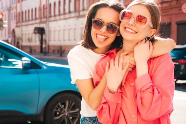 Son moda beyaz tişörtlü ve kot pantolonlu iki güzel, gülümseyen yeniyetme bayan sokak arka planında poz veren kaygısız kadınlar. Pozitif modeller eğleniyor, sarılıyor ve deliriyor.