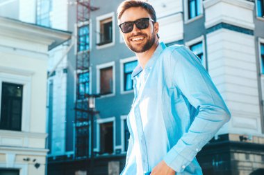 Yakışıklı, güler yüzlü, stil sahibi, lamberseksüel bir modelin portresi. Mavi gömlekli modern bir adam. Moda erkeği güneş gözlükleriyle gökdelenin yanında poz veriyor. Gün batımında dışarıda. 