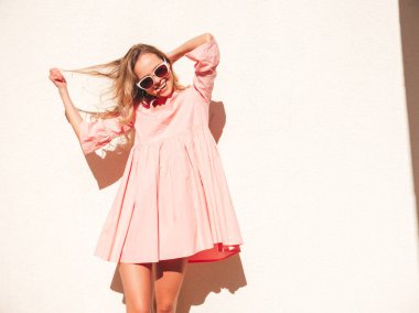 Son moda pembe elbiseli güzel, gülümseyen hippi kadın. Günbatımında sokakta beyaz duvarın yanında poz veren kaygısız seksi kadın. Evrak çantalı olumlu model. Mutlu ve neşeli.