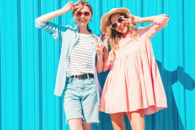 Son moda yaz kıyafetleri ve elbiseleri içinde iki güzel gülümseyen yeniyetme kadın. Mavi duvarın yanındaki sokakta poz veren kaygısız kadınlar. Gün batımında eğlenen, sarılan ve deliren pozitif modeller.