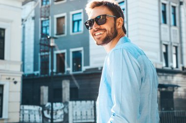 Yakışıklı, güler yüzlü, stil sahibi, lamberseksüel bir modelin portresi. Mavi gömlekli modern bir adam. Moda erkeği güneş gözlükleriyle gökdelenin yanında poz veriyor. Gün batımında dışarıda. 