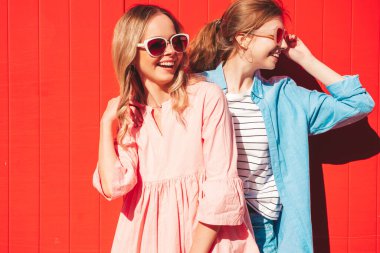 Son moda yaz kıyafetleri içinde iki güzel, gülümseyen yeniyetme kadın. Kırmızı renkli duvarın yanında sokakta poz veren kaygısız kadınlar. Pozitif modeller eğleniyor ve sarılıyorlar. Neşeli ve mutlu
