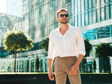 Yakışıklı, gülümseyen stil sahibi Lambersex modelinin portresi. Beyaz gömlek giymiş modern bir adam. Moda erkeği, güneş gözlüklü gökdelenlerin yanında sokak arkasında poz veriyor.