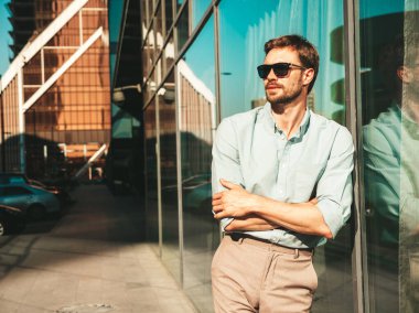 Yakışıklı, gülümseyen stil sahibi Lambersex modelinin portresi. Beyaz gömlek giymiş modern bir adam. Moda erkek, güneş gözlüklü gökdelenlerin yanında sokak arkasında poz veriyor. Çapraz kollar