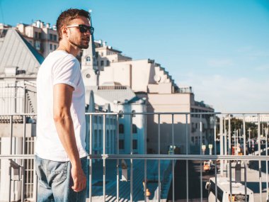 Yakışıklı, kendine güvenen hipster Lambersex modelinin portresi. Beyaz tişört giyen şık bir adam. Moda erkeği mavi gökyüzünün arkasında güneş gözlükleriyle sokak arkasında poz veriyor.