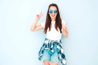 Yeni moda hippi kıyafetleri içinde gülümseyen güzel bir kadın. Stüdyoda açık mavi duvarın yanında poz veren kaygısız seksi kadın. Pozitif model içeride eğleniyor. Neşeli ve mutlu. Barış işareti nasıl?