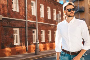 Yakışıklı, kendine güvenen, stil sahibi, Lambersex mankeni portresi. Beyaz gömlek giymiş modern bir adam. Moda erkek, güneş gözlüklü gökdelenlerin yanında sokak arkasında poz veriyor. Gün batımında dışarıda. 