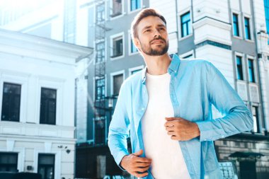 Yakışıklı, güler yüzlü, stil sahibi, lamberseksüel bir modelin portresi. Mavi gömlekli modern bir adam. Moda erkeği, arka planda gökdelenin yanında poz veriyor. Gün batımında dışarıda. 