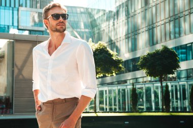Yakışıklı, kendine güvenen, stil sahibi, Lambersex mankeni portresi. Beyaz gömlek giymiş modern bir adam. Moda erkeği, güneş gözlüklü gökdelenlerin yanında sokak arkasında poz veriyor.
