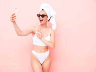 Beyaz iç çamaşırlı, gülümseyen genç bir kadın. İç çamaşırları ve kafasında havluyla stüdyoda pembe duvar pozu veren seksi tasasız manken. Pozitif ve mutlu bir kadın güneş gözlükleriyle sabahın tadını çıkarıyor. Selfie çekiyor.