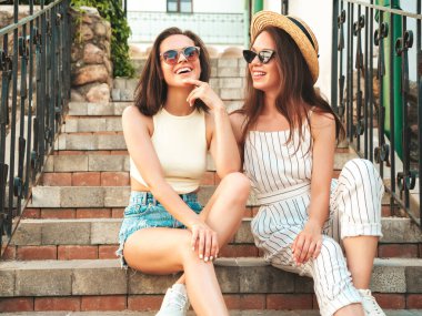 Son moda yaz kıyafetleri içinde iki güzel gülümseyen yeniyetme kadın. Sokakta şapkalı kayla poz veren kaygısız kadınlar. Pozitif saf modeller gün batımında eğleniyorlar. Merdivenlerde oturuyor.