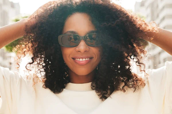 Beautiful black woman with afro curls hairstyle.Smiling hipster model in white t-shirt. Sexy carefree female posing on the street background. Cheerful and happy in sunglasses