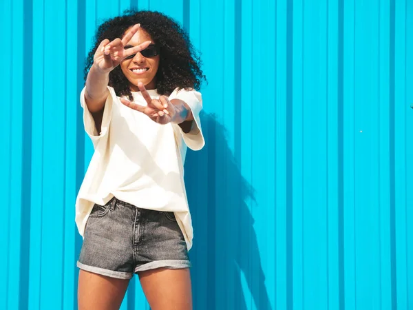 Beautiful black woman with afro curls hairstyle.Smiling hipster model in white t-shirt. Sexy carefree female posing in the street near blue wall in sunglasses. Cheerful and happy.Shows peace sign