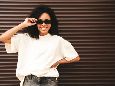 Saçları kıvırcık, güzel siyah bir kadın. Beyaz tişörtlü, hippi bir model. Sokakta kahverengi duvarın yanında poz veren kaygısız seksi kadın. Neşeli ve mutlu, güneş gözlükleriyle