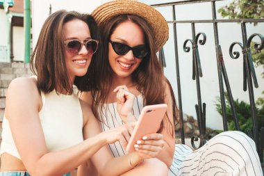 Son moda yaz kıyafetleri içinde iki güzel gülümseyen yeniyetme kadın. Sokakta poz veren kaygısız kadınlar. Akıllı telefon uygulamaları kullanan, ekrana bakan pozitif modeller.