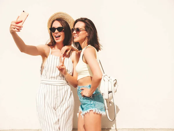 流行の夏服の2人の若い美しい笑顔のヒップスター女性 帽子の白い壁の近くの通りにポーズ性的屈託のない女性 正の純粋なモデルは 夕日を楽しみます セルフィーを取ります — ストック写真
