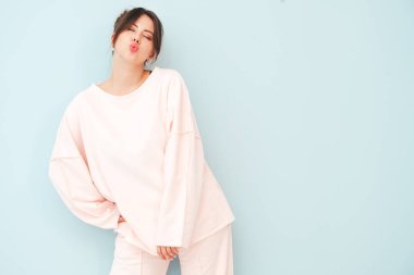 Son moda pembe giysiler içinde gülümseyen güzel bir kadının portresi. Stüdyoda açık mavi duvarın yanında oturan kaygısız seksi kadın. Pozitif model içeride eğleniyor. Ördek suratı yapar