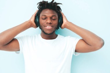 Yakışıklı, güler yüzlü, lamberseksüel model. Şık giyinmiş, traşsız Afrikalı bir erkek. Stüdyoda duvarın yanında saç stili saçı olan, kablosuz kulaklıkla müzik dinleyen moda bir erkek.