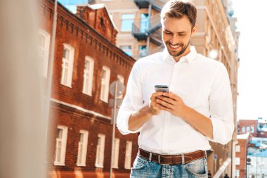 Yakışıklı, güler yüzlü, stil sahibi, lamberseksüel model. Beyaz gömlekli, modern bir adam. Moda erkeği sokakta poz veriyor. Gün batımında dışarıda. Akıllı telefon uygulamaları kullanarak. Cep telefonu tutuluyor