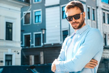 Yakışıklı, kendine güvenen, havalı, havalı, lamberseksüel model. Mavi gömlekli, modern bir adam. Moda erkeği güneş gözlükleriyle gökdelenin yanında poz veriyor. Gün batımında dışarıda. Çapraz kollar.