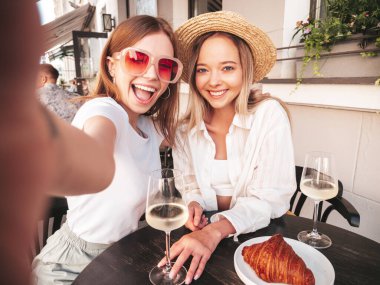 Son moda yaz kıyafetleri içinde gülümseyen iki genç bayan. Sokakta Veranda Kafe 'de poz veren kaygısız kadınlar. Beyaz şarap içen, kruvasan yiyen, Pov selfie çeken kadınlar.