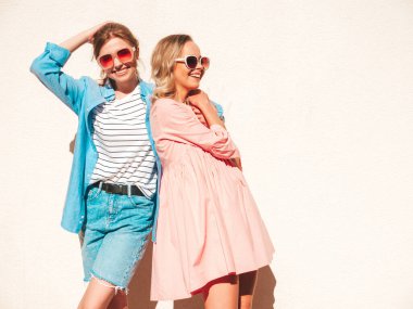 Son moda yaz kıyafetleri ve elbiseleri içinde iki güzel, gülümseyen hippi kadın. Sokakta beyaz duvarın yanında poz veren kaygısız kadınlar. Pozitif saf modeller eğleniyor, sarılıyor ve deliriyor.