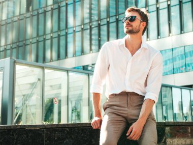 Yakışıklı, kendine güvenen, stil sahibi, Lambersex mankeni portresi. Beyaz gömlek giymiş modern bir adam. Moda erkeği, güneş gözlüklü gökdelenlerin yanında sokak arkasında poz veriyor.