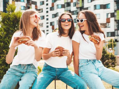Üç güzel, gülümseyen yeniyetme kadın yazın moda olan aynı kıyafetleri giyiyor. Sokakta poz veren kaygısız seksi kadınlar. Pozitif modeller güneş gözlükleriyle eğleniyor. Sulu burger yiyip,