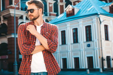 Yakışıklı, şık, havalı, havalı, lamberseksüel model. Kırmızı kareli gömlek giymiş modern bir adam. Moda erkeği güneş gözlükleriyle gökdelenin yanında poz veriyor. Gün batımında dışarıda. 