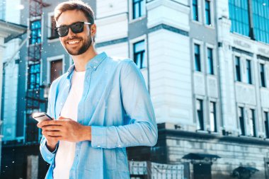 Yakışıklı, güler yüzlü, stil sahibi, lamberseksüel model. Mavi gömlekli, modern bir adam. Sokakta güneş gözlükleriyle poz veren modacı erkek. Gün batımında dışarıda. Akıllı telefon uygulamaları kullanıyor. SMS yazıyor.