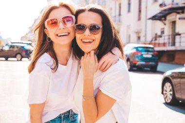 Son moda beyaz tişörtlü ve kot pantolonlu iki güzel, gülümseyen yeniyetme bayan sokak arka planında poz veren kaygısız kadınlar. Pozitif modeller eğleniyor, sarılıyor ve deliriyor.