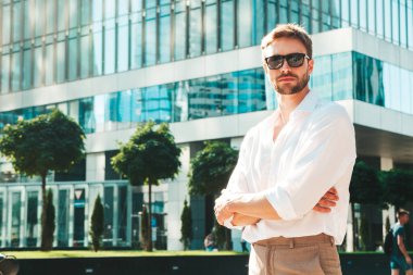 Yakışıklı, kendine güvenen, stil sahibi, Lambersex mankeni portresi. Beyaz gömlek giymiş modern bir adam. Moda erkek, güneş gözlüklü gökdelenlerin yanında sokak arkasında poz veriyor. Çapraz kollar