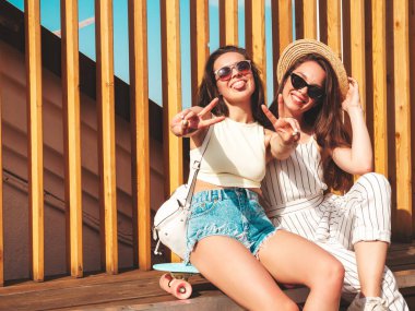 Son moda yaz kıyafetleri içinde iki güzel gülümseyen yeniyetme kadın. Sokakta şapkalı kayla poz veren kaygısız kadınlar. Pozitif saf modeller gün batımında eğleniyorlar, sarılıyorlar ve deliriyorlar.