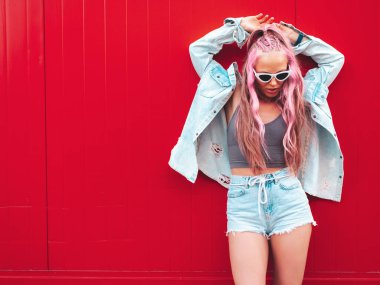 Genç, güzel, gülümseyen bir hippi kadın, son moda yaz kot şortuyla, pembe saç stiliyle. Sokakta kırmızı duvarın yanında poz veren kaygısız seksi bir model. Güneş gözlüklü pozitif model.