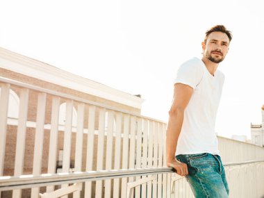 Yakışıklı, gülümseyen hipster Lambersex modelinin portresi. Beyaz tişört ve kot giyen şık bir adam. Moda erkeği, plajdaki çitlerin yanında sokak arkasında poz veriyor.