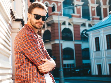 Yakışıklı, şık, havalı, havalı, lamberseksüel model. Kırmızı kareli gömlek giymiş modern bir adam. Moda erkeği güneş gözlükleriyle gökdelenin yanında poz veriyor. Gün batımında dışarıda. 