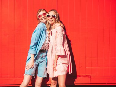 Son moda yaz kıyafetleri içinde iki güzel, gülümseyen yeniyetme kadın. Kırmızı renkli duvarın yanında sokakta poz veren kaygısız kadınlar. Pozitif modeller eğleniyor ve sarılıyorlar. Neşeli ve mutlu
