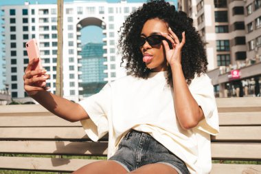 Saçları kıvırcık, güzel siyah bir kadın. Beyaz tişörtlü, hippi bir model. Sokakta güneş gözlükleriyle poz veren kaygısız seksi kadın. Neşeli ve mutlu. Selfie çekiyoruz.